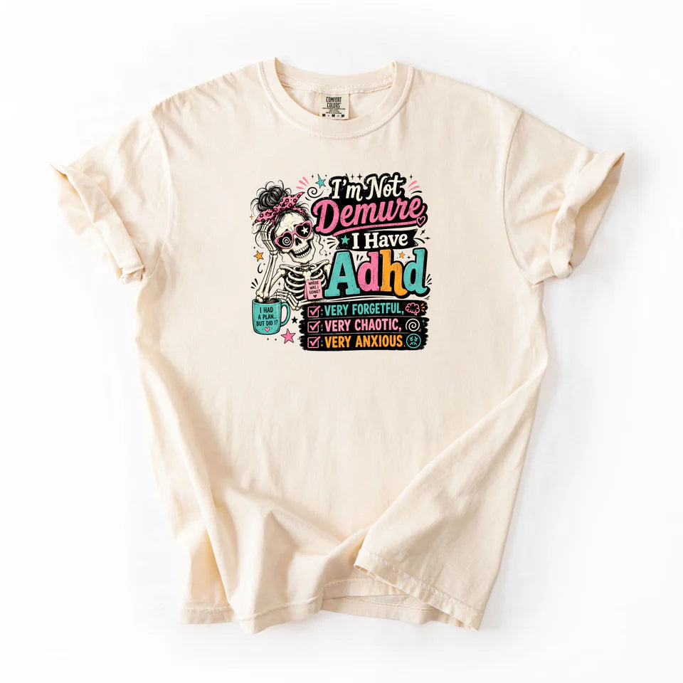 Ivory Comfort Colors flat lay tee with I'm Not Demure I Have ADHD graphic featuring messy bun skeleton with sunglasses and coffee mug very forgetful very chaotic very anxious