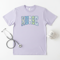 Flat lay of lavender nurse patchwork graphic tee on white background styled with stethoscope and coffee mug